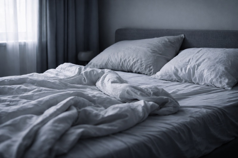 Lit défait en plein jour avec rideaux fermés, illustrant la fatigue et l’épuisement physique et mental.