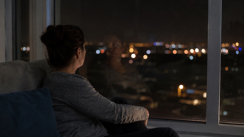 isolement-nocturne-sp Reflet flou d’une femme dans une fenêtre la nuit, avec des lumières de la ville en arrière-plan, illustrant l’isolement émotionnel vécu par certaines personnes atteintes de sclérose en plaques.